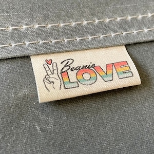May include: A white fabric label with the text "Beanie LOVE" in rainbow letters. A peace sign with a heart is on the left side of the label.