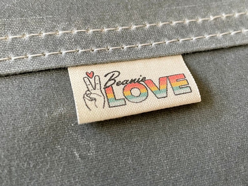May include: A white fabric label with the text "Beanie LOVE" in rainbow letters. A peace sign with a heart is on the left side of the label.