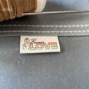 May include: A white fabric label with the text "Beanie's LOVE" in rainbow colors. A peace sign with a small red heart is above the text.