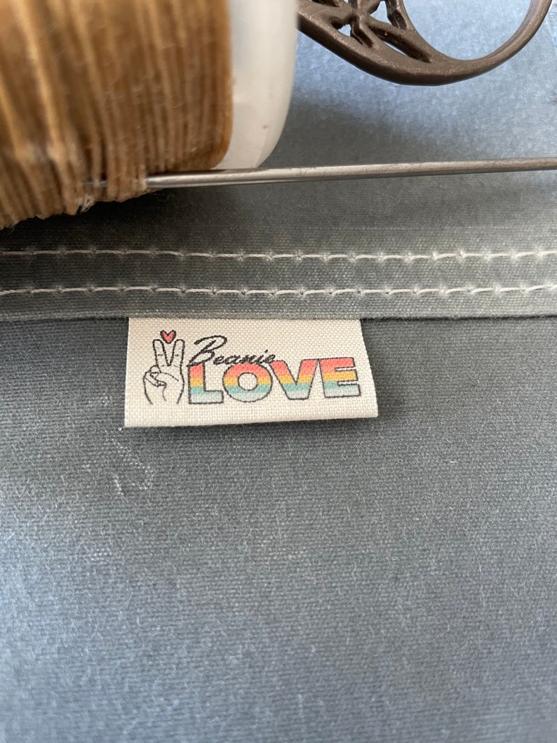 May include: A white fabric label with the text "Beanie's LOVE" in rainbow colors. A peace sign with a small red heart is above the text.