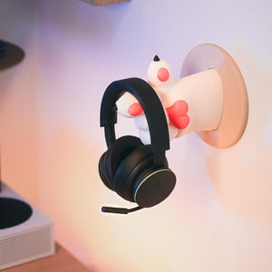Può includere: Un supporto per cuffie a forma di zampa di gatto bianco e rosa montato su una parete con un paio di cuffie nere appese.