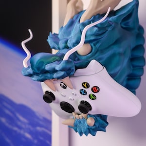 Könnte beinhalten: Ein Wandhalter in Form eines türkisblauen Drachenkopfes mit einem weißen Xbox-Controller im Maul. Der Drache hat weiße Hörner und eine strukturierte Oberfläche.