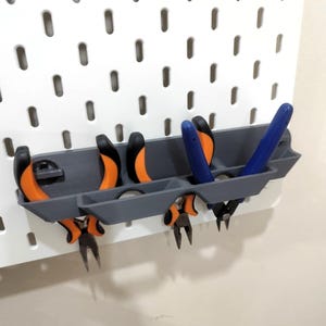 May include: A grey pegboard organiser holding pliers with orange and black handles, and blue handles. The organiser is mounted on a white pegboard with evenly spaced holes.
