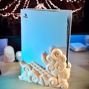 May include: A white PlayStation 5 console with a cloud-shaped stand. The stand is made of a light beige plastic and features a swirling cloud design.