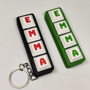 Könnte beinhalten: Zwei Schlüsselanhänger mit dem Namen "EMMA", geschrieben mit einzelnen Tastaturtasten. Der schwarze Schlüsselanhänger hat rote Buchstaben, der grüne hat grüne Buchstaben. Ein silberner Schlüsselring ist am schwarzen Schlüsselanhänger befestigt.