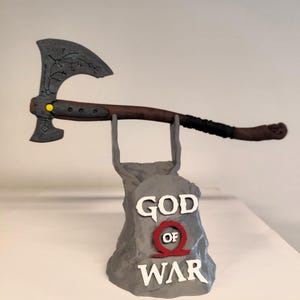 Puede incluir: Un soporte de exposición impreso en 3D gris con las palabras "God of War" en él. Un hacha negra y plateada se muestra en el soporte.