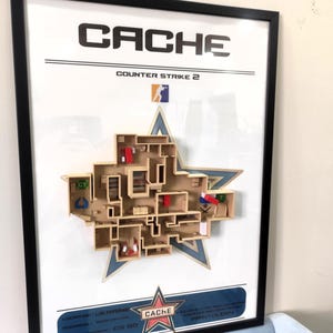 May include: A framed 3D wall art piece featuring the Cache map from Counter Strike 2. The display includes a tan-colored architectural layout of the map set against a blue star background, with text labels including Cache, Counter Strike 2, and Location: Ukraine.