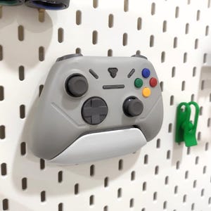 Pode incluir: Um controle de videogame cinza e branco com botões coloridos e um direcional está montado em uma parede perfurada branca. O controle tem um logotipo e dois joysticks pretos. Um clipe verde está à direita.