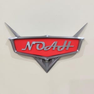 Może przedstawiać: Wydrukowany w 3D znak z napisem "NOAH" w srebrnych literach na czerwonym tle. Znak ma stylizowany, motoryzacyjny design ze srebrną obwódką i spiczastymi akcentami. Zamontowany na kremowej powierzchni.