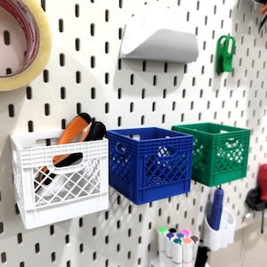 Puede incluir: Un organizador de pared de tablero perforado blanco con tres pequeñas cajas de almacenamiento de plástico en blanco, azul y verde. Las cajas están montadas en el tablero, con la caja blanca conteniendo herramientas naranjas y negras.