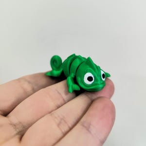 Puede incluir: Una figura de camaleón verde articulada con ojos blancos y negros. El camaleón de juguete está hecho de plástico y tiene un cuerpo segmentado, lo que permite el movimiento. La figura se sostiene en una mano.