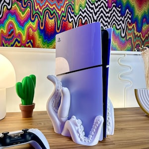 May include: A white octopus-shaped stand holding a purple PlayStation 5 console. The console is standing vertically and has the PlayStation logo on the front. The stand is on a wooden surface.