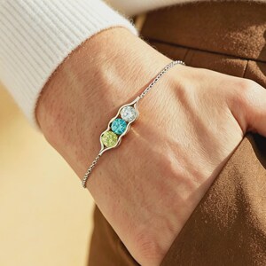 May include: A silver bracelet featuring a pea pod design with three colorful gemstones. The gemstones are white, blue, and green. The bracelet is worn on a wrist, and the background is a neutral color.