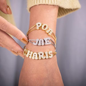 May include: Three personalized name bracelets. The top bracelet is gold-toned with the name "POR" spelled out in sparkling crystals. The middle bracelet is silver-toned with the name "JAE". The bottom bracelet is gold-toned with the name "CHARIS".