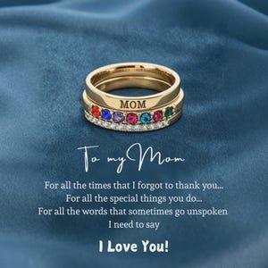 May include: Gold-toned rings stacked together, one engraved with "MOM" and adorned with colorful gemstones. The rings sit on a blue surface. The text "To my Mom" and a loving message are also visible.