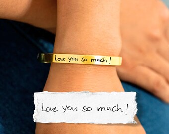 May include: A gold-toned metal cuff bracelet featuring the engraved black text "Love you so much!" in a handwritten script style. The bracelet is displayed alongside a white paper scrap showing the same handwritten text "Love you so much!"