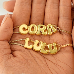 Op de afbeelding: Twee gouden kettingen met de namen "CORA" en "LULU" gespeld in bubbelletters.