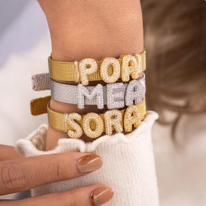May include: Three personalized bracelets stacked on a wrist. The top and bottom bracelets are gold-toned mesh with diamond-studded letter charms spelling "POA" and "SORA". The middle bracelet is silver-toned with "MEA" charms.