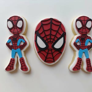 May include: Three Spider-Man themed cookies. Two cookies are shaped like the character, with red and black spider-web design, blue accents, and white eyes. The third cookie is a Spider-Man mask with a red spider-web design and white eyes.