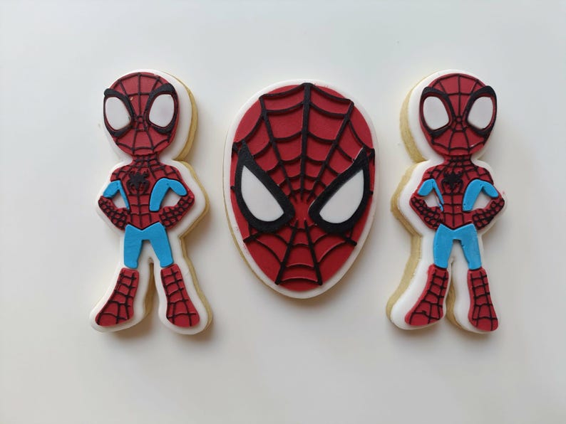 May include: Three Spider-Man themed cookies. Two cookies are shaped like the character, with red and black spider-web design, blue accents, and white eyes. The third cookie is a Spider-Man mask with a red spider-web design and white eyes.