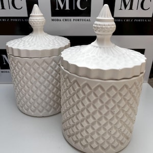 Pode incluir: Dois frascos de vela de cerâmica branca com padrões em forma de diamante e tampas ornamentadas. Os frascos estão sobre uma superfície branca. O texto "MIC MODA CRUZ PORTUGAL" é visível no fundo.