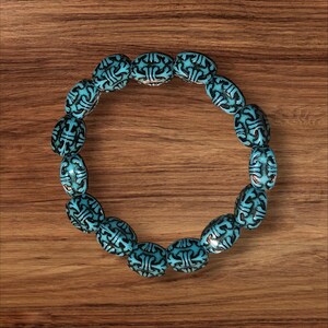 May include: A turquoise beaded bracelet with black etched designs. The oval beads are arranged in a circular shape, showcasing a detailed pattern. The bracelet is set against a wooden background.