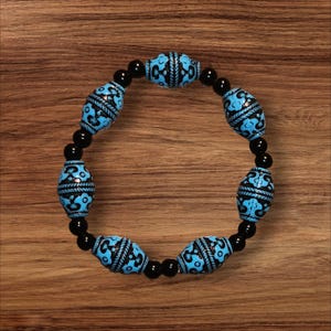 May include: A beaded bracelet featuring alternating black and turquoise beads. The turquoise beads are cylindrical with a black, patterned design. The bracelet is strung on a black elastic cord and rests on a wooden surface.
