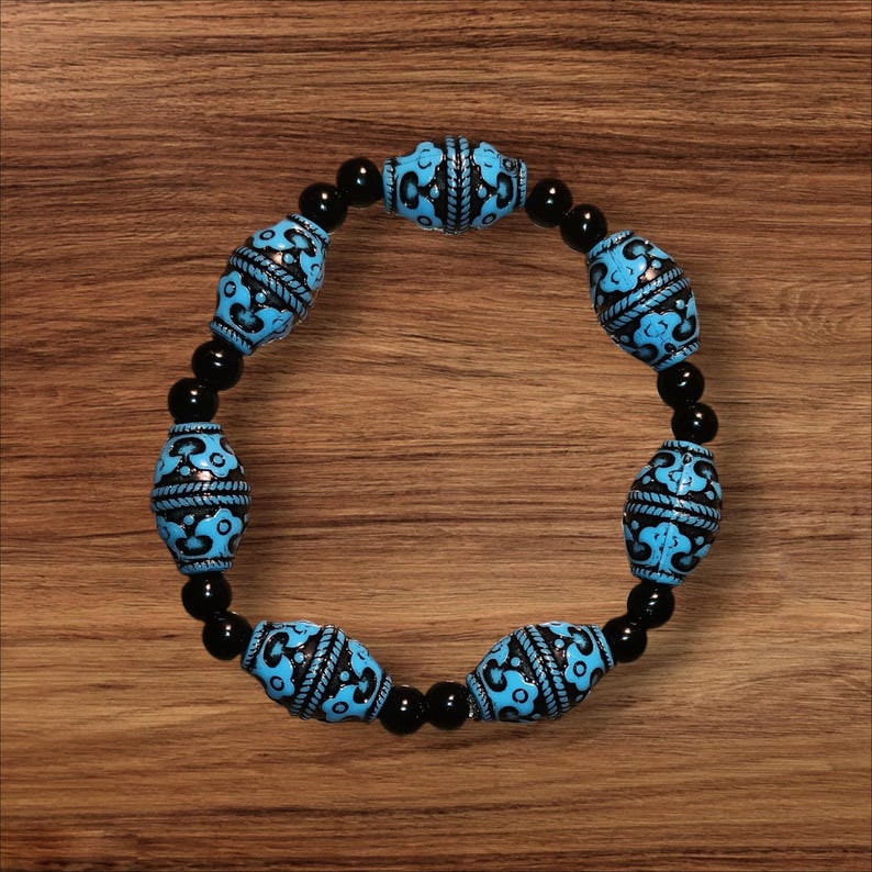 May include: A beaded bracelet featuring alternating black and turquoise beads. The turquoise beads are cylindrical with a black, patterned design. The bracelet is strung on a black elastic cord and rests on a wooden surface.
