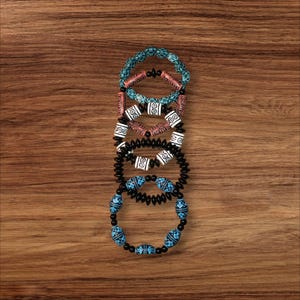 May include: Five beaded bracelets in various colors and designs are stacked on a wooden surface. The bracelets feature turquoise, coral, black, and white beads with intricate patterns. The bracelets are a mix of round and oblong beads.