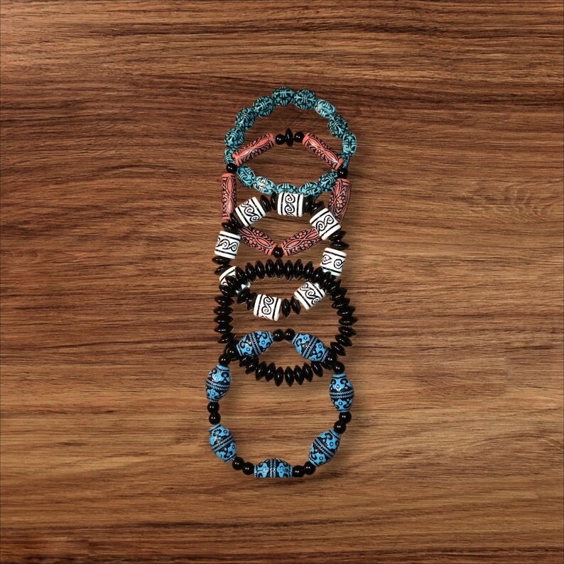 May include: Five beaded bracelets in various colors and designs are stacked on a wooden surface. The bracelets feature turquoise, coral, black, and white beads with intricate patterns. The bracelets are a mix of round and oblong beads.