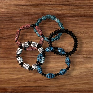 May include: Four beaded bracelets in various colors and designs are displayed on a wooden surface. The bracelets feature black, white, blue, and pink beads with intricate patterns. One bracelet is a solid black coil.