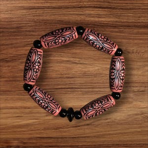 May include: A beaded bracelet featuring pink, barrel-shaped beads with black etched floral designs. The bracelet is strung with black beads and is displayed on a wooden surface.