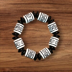 May include: A black and white beaded bracelet. The bracelet features cylindrical white beads with a black swirl design, interspersed with smaller, black, oval-shaped beads. The bracelet is displayed on a wooden surface.