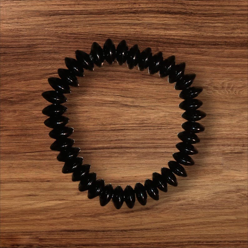 May include: A black, flexible, segmented ring-shaped object. The segments are oval-shaped and glossy, arranged in a circular pattern. The object is set against a wooden background.