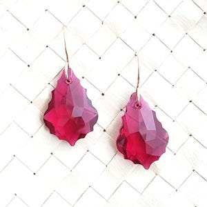 May include: A pair of drop earrings featuring faceted, deep magenta-colored crystal pendants with a scalloped edge. Each crystal is suspended from a thin, curved gold-toned metal wire hook, creating a dangling design that catches the light.
