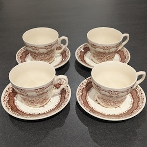 May include: A set of four cream-coloured ceramic teacups and matching saucers, each decorated with a brown floral pattern and a detailed border design. The cups feature a curved handle and a wide rim, while the saucers have a matching brown rim.