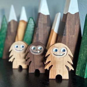 May include: Three wooden toy monsters with painted faces and smiling expressions. The monsters are standing in front of a background of wooden mountains.