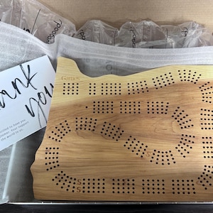 May include: A wooden game board shaped like the state of Oregon. The board has a light brown color and is made of wood. The board has a variety of holes and dots on it. The board is in a box with a thank you note.