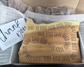 May include: A wooden game board shaped like the state of Oregon. The board has a light brown color and is made of wood. The board has a variety of holes and dots on it. The board is in a box with a thank you note.
