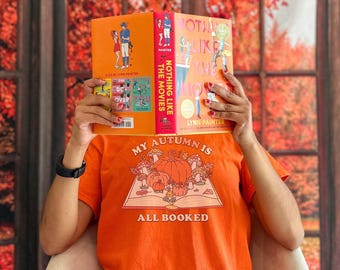 May include: An orange t-shirt featuring a graphic of an open book with pumpkins and mushrooms, with the text "MY AUTUMN IS ALL BOOKED". The shirt is worn by someone holding a book titled "NOTHING LIKE THE MOVIES" by Lynn Painter in front of their face.