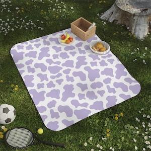 May include: A picnic blanket with a white background and a repeating pattern of lavender-coloured cow spots. A football, tennis racket, and food items are on the grass next to the blanket. A small basket and a plate of fruit are on the blanket.