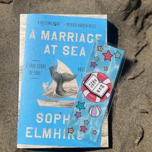 Pode incluir: Um livro intitulado "A Marriage at Sea" com uma capa azul clara e um marcador. O marcador tem um tema de praia com estrelas, uma bola de praia e o texto "My only plan is to read all day."