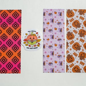 May include: Three rectangular bookmarks and one die-cut sticker. One bookmark has a black geometric pattern on an orange-to-pink gradient. Another is light purple with books and mugs. The third has pumpkins and the text "BOO".