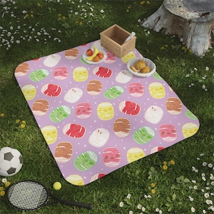 Può includere: Una coperta da picnic lavanda con un motivo di pasticcini illustrati colorati. Sull'erba verde, ci sono un pallone da calcio, una racchetta da tennis e un piccolo cestino di cibo.