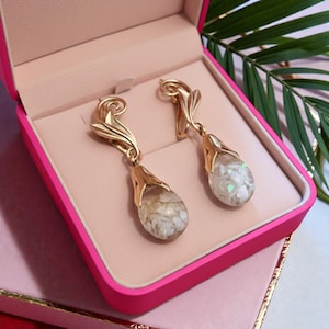 May include: Gold-toned dangle earrings with a floral design. Each earring features a teardrop-shaped, clear resin pendant filled with white and beige stones. The earrings are displayed in a pink jewelry box.