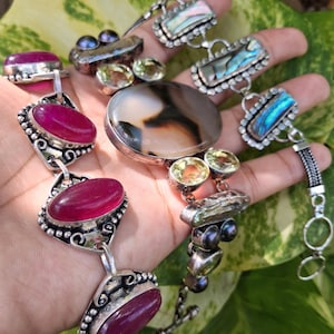 May include: Three silver bracelets featuring distinct gemstones. One bracelet has oval magenta stones, another displays a large central agate with yellow and dark stones, and the third showcases rectangular iridescent abalone shell segments.