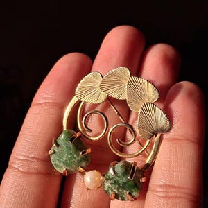 Puede incluir: Broche vintage dorado con detalles de piedras verdes y una perla. El broche tiene un diseño de hojas con detalles intrincados y un cierre de alfiler. El broche mide aproximadamente 5 cm de diámetro.