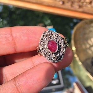 Puede incluir: Anillo de plata con una piedra preciosa roja ovalada. El anillo presenta un diseño de filigrana ornamentado alrededor de la piedra, con detalles dorados. El anillo está sostenido por una mano.