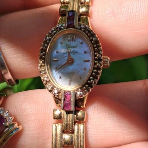 May include: A gold-tone wristwatch with an oval-shaped mother-of-pearl dial, featuring the text Gerard Pullipe. The bezel is accented with small clear stones and two pink stones at the top and bottom. It has a link bracelet and quartz movement.