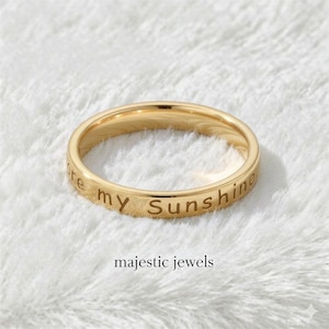 以下が含まれることがあります： 「You are my Sunshine」の文字が刻まれたゴールドカラーのリング。リングは柔らかく白いテクスチャの背景に配置されています。「majestic jewels」の文字が下にあります。